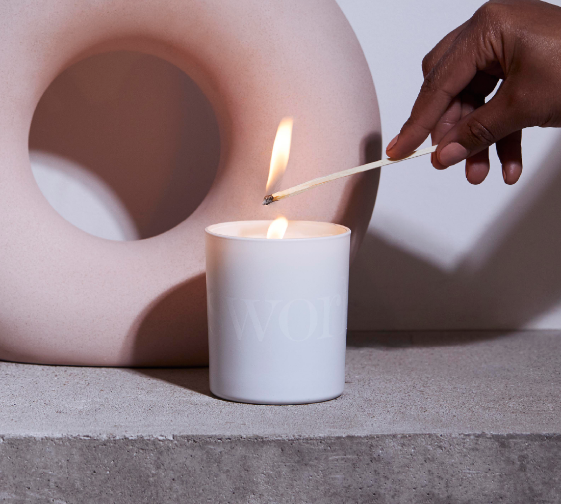 A white candle is lit with a match held by a hand, with a large pale pink object in the background on a gray surface.