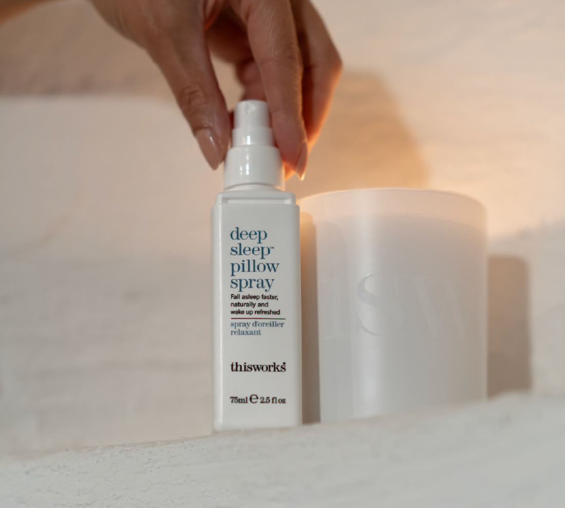 A bottle of thisworks Deep Sleep Pillow Spray stands on a light surface, with a candle blurred in the background.