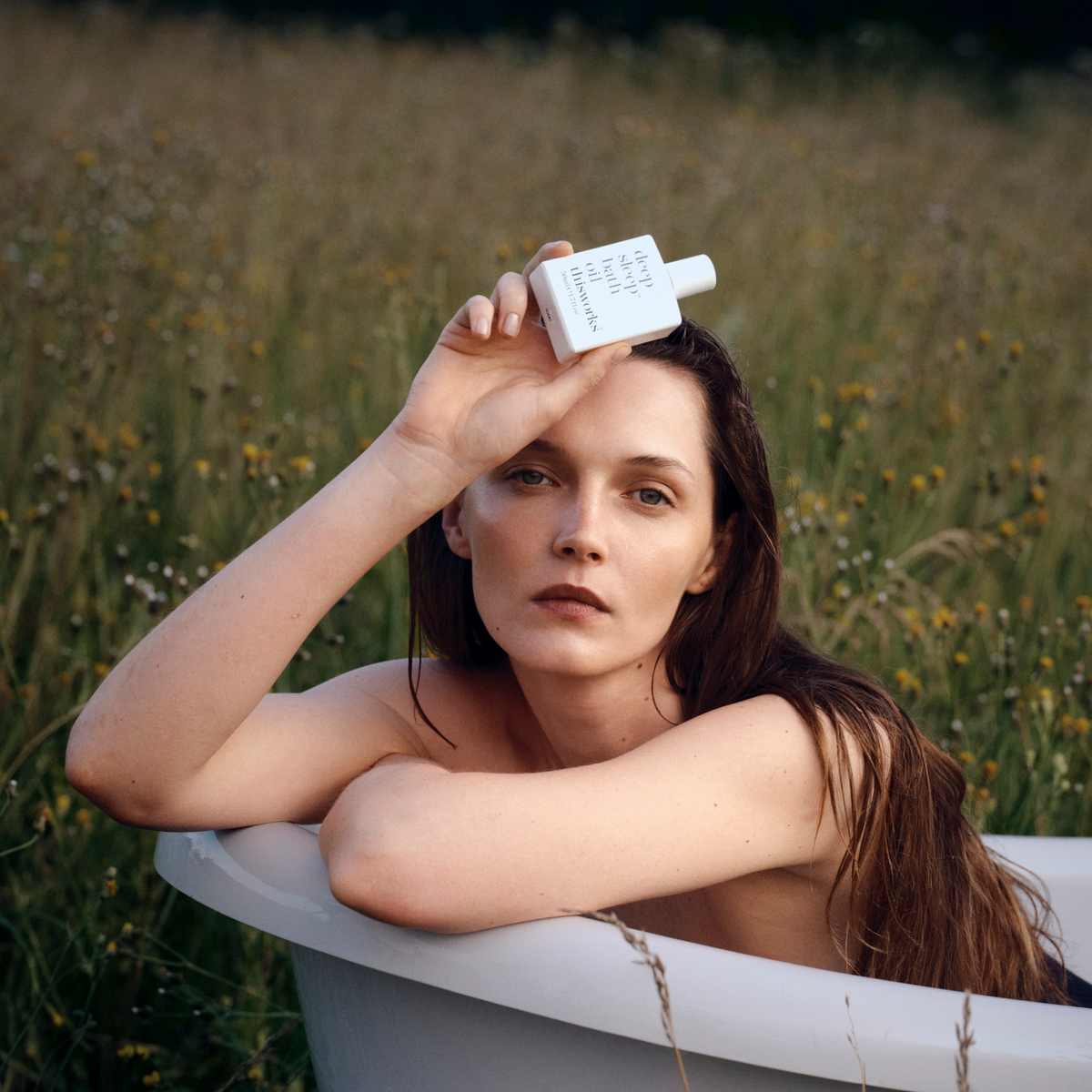 Woman resting in a white bathtub set in a wildflower field, holding This Works Deep Sleep Bath Oil bottle above her head.