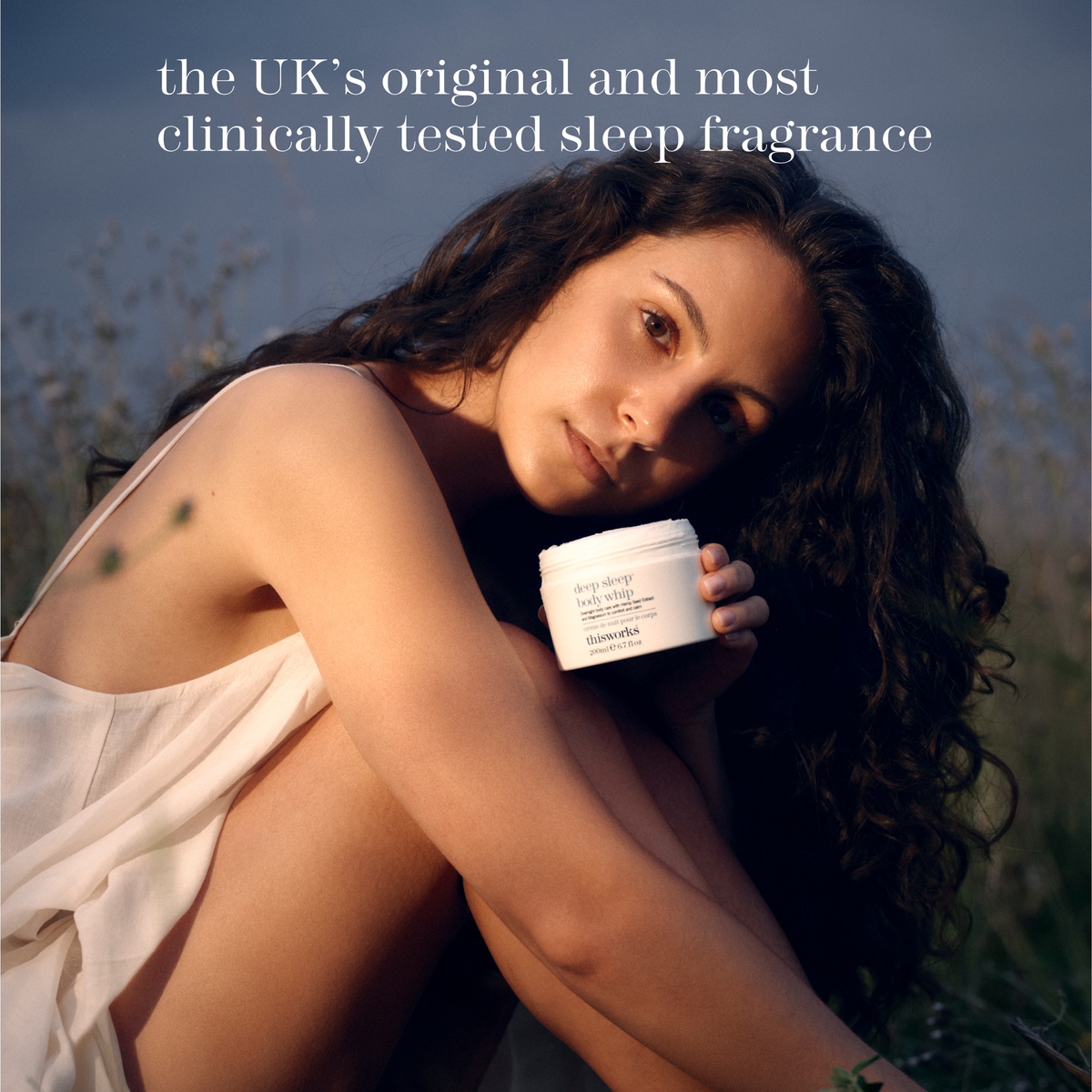 A woman outdoors in warm golden hour light holds a white jar of deep sleep body whip.