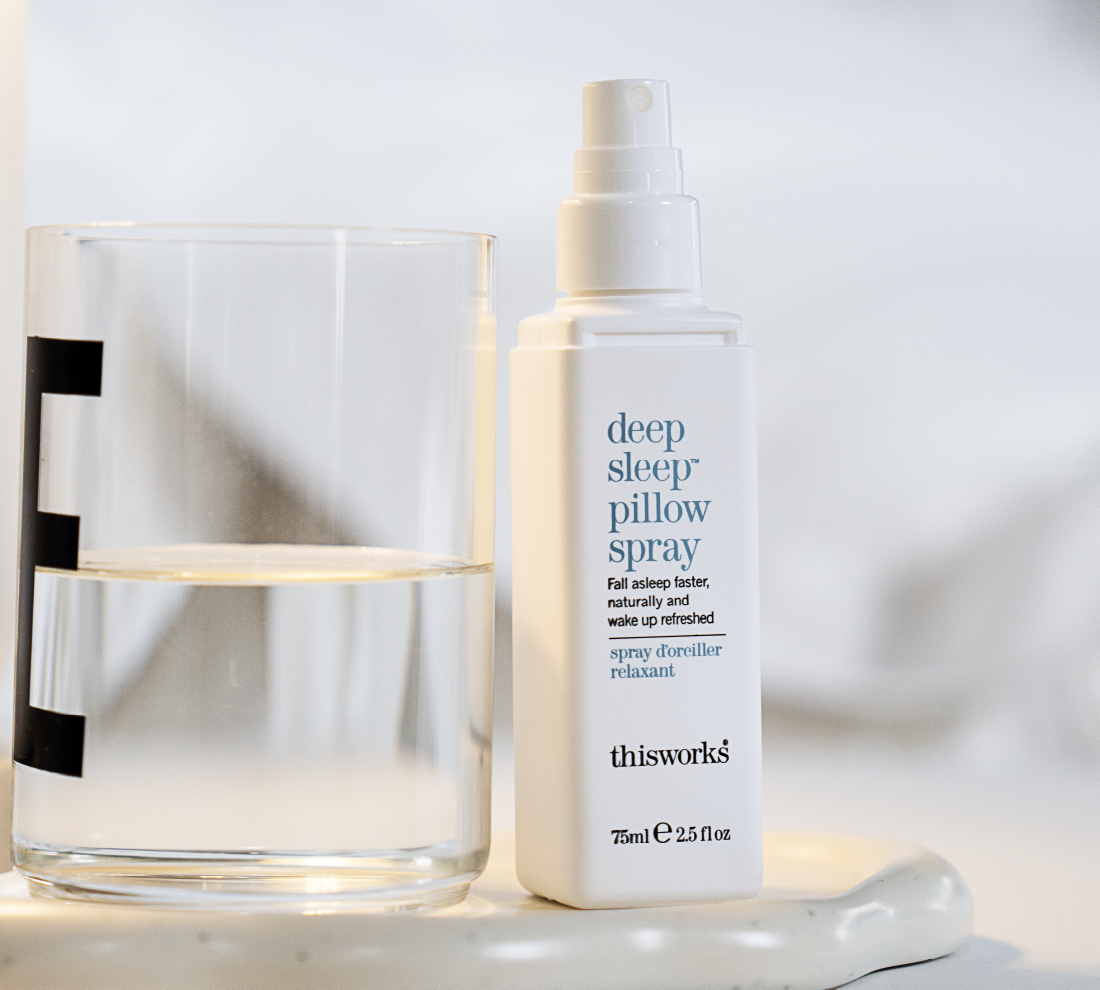 A bottle of thisworks Deep Sleep Pillow Spray standing next to a clear glass of water on a white marble surface.