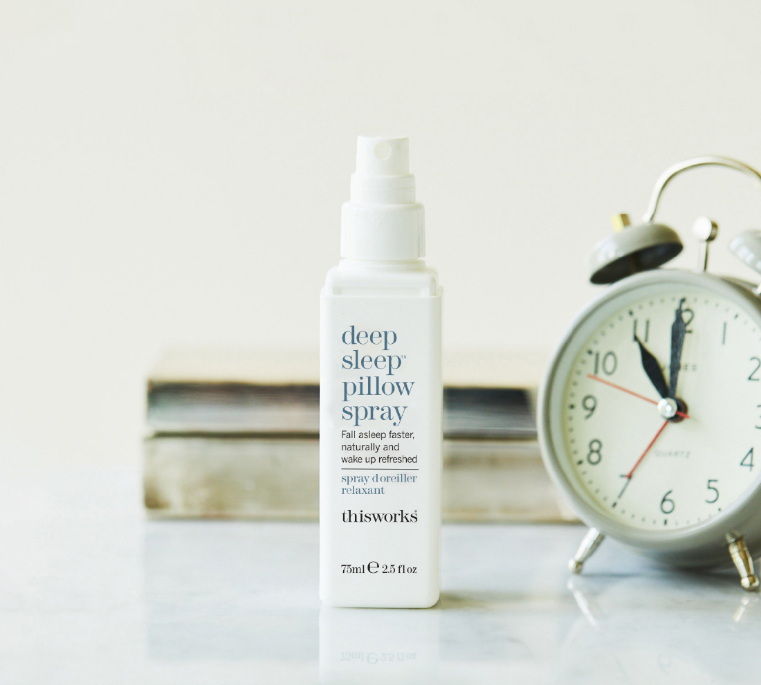 A bottle of thisworks Deep Sleep Pillow Spray stands on a light surface, with a silver alarm clock and stacked books blurred in the background.