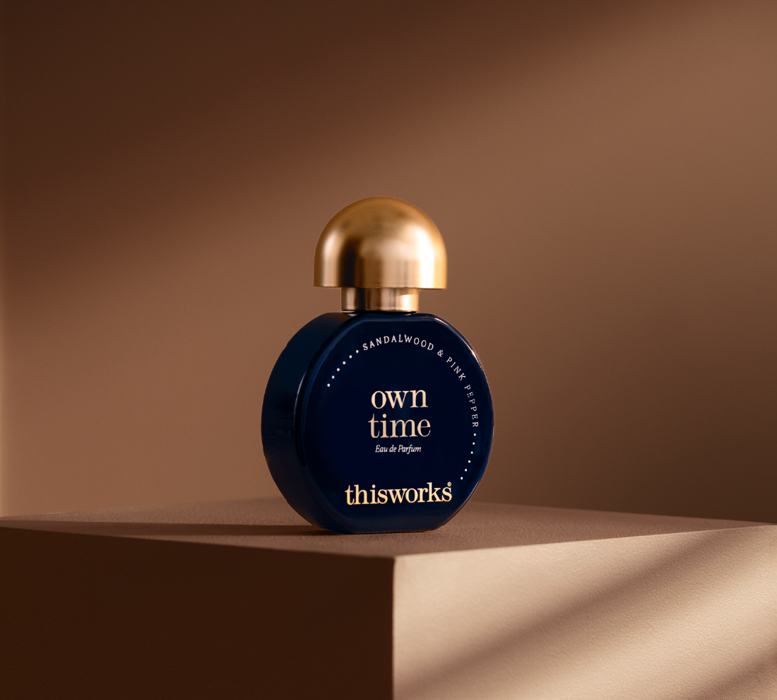 A dark blue spherical perfume bottle with a metallic gold cap, labeled 'own time' by thisworks, displayed on a matching brown pedestal against a warm brown background.