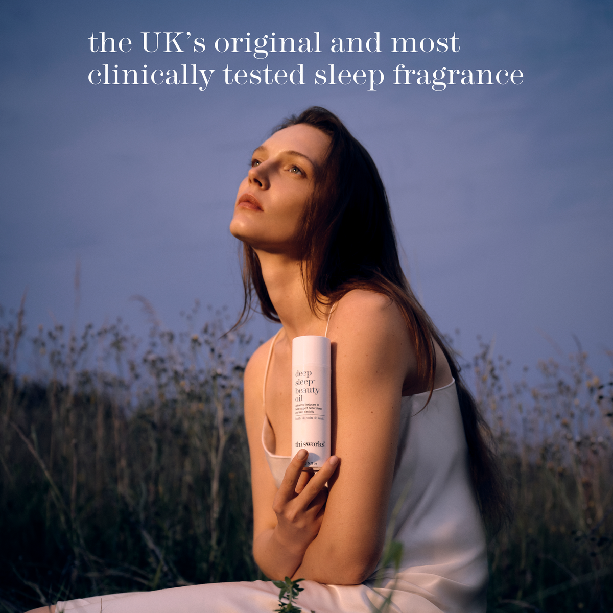A woman in a white dress sits outdoors in a field at dusk, holding a white bottle labelled deep sleep beauty oil. 'the UK's original and most clinically tested sleep fragrance.